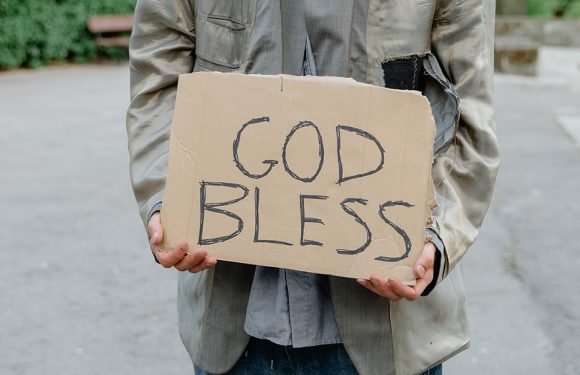 Person outside on a sidewalk holding a handwritten cardboard sign that reads "God Bless".