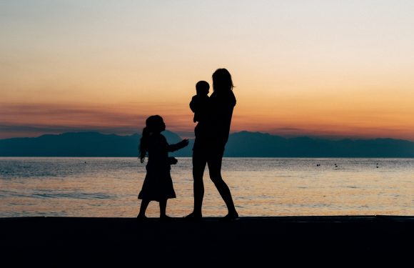 Silhouette of a woman holding infant with young child standing next to them on the beach in front of a sunset.