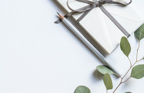 white flat lay of green leaves, silver sharpened pencil and ribbon wrapped envelopes