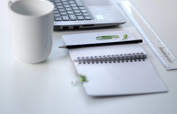 open laptop with coffee cup, notebook, ruler, thumbtack and paperclip on top of a white desk