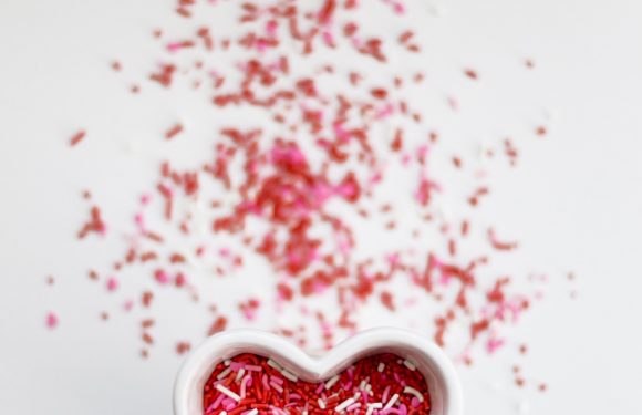 White background with small white heart-shaped bowl filled with red, pink and white sprinkles and some of the sprinkles spilled around the bowl