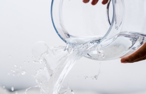 Hands holding a clear glass jar pouring water into another glass container with it overflowing and spilling all over.