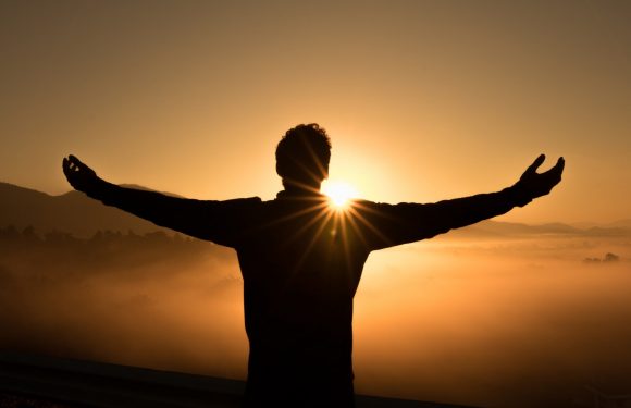 Silhouette of man with arms stretched out to the sides in front of a sunrise over misty mountains