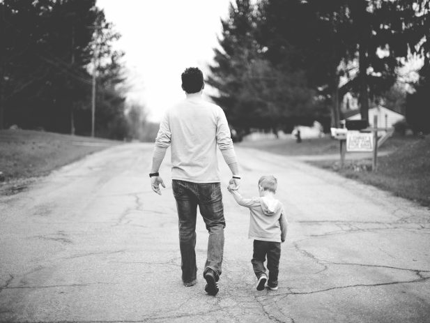 Black and white photo of father and young son holding hands and walking away down a paved residential road.