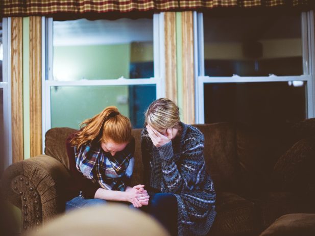 Two people with heads down together on a couch. One lady has her head in her hand and holding the hands of the other lady in prayer.