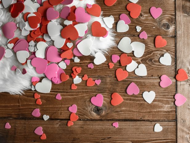 Wooden table with red, pink and white cutout paper hearts and white fuzzy fabric laid over the top.