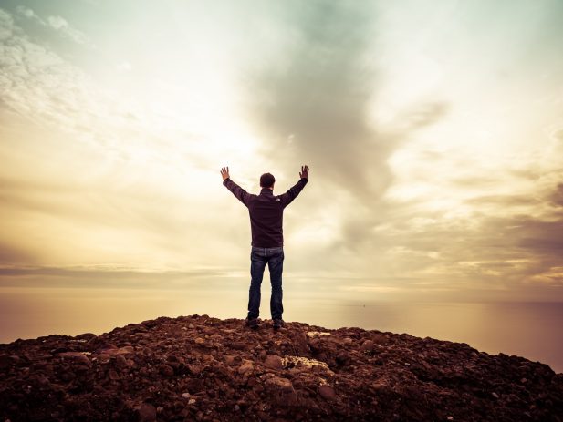 Man standing under orange sky on a rocky hill with arms outstretched toward the sky.