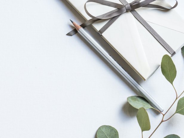 white flat lay of green leaves, silver sharpened pencil and ribbon wrapped envelopes