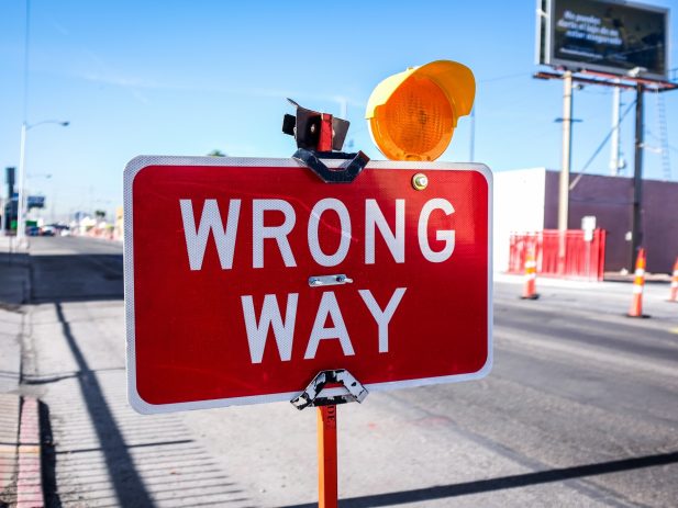 Photo of a construction sign saying "wrong way" with a flashing light on it, traffic cones and street in the background.