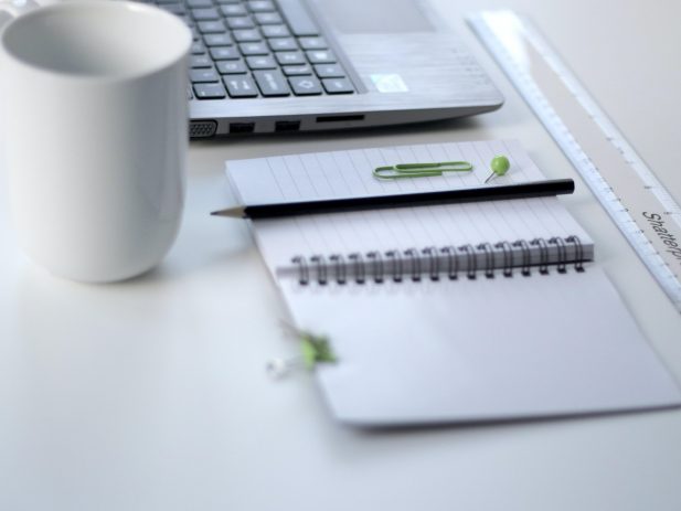 open laptop with coffee cup, notebook, ruler, thumbtack and paperclip on top of a white desk