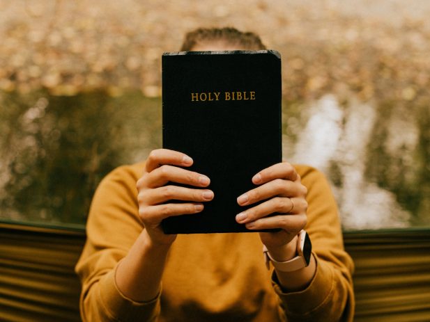 Girl in a yellow sweater holding a bible out in front of her face.