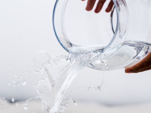 Hands holding a clear glass jar pouring water into another glass container with it overflowing and spilling all over.