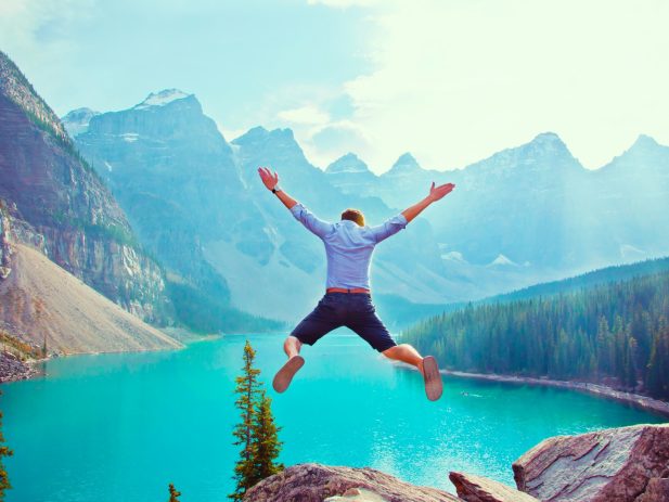 Person jumping up in the air spread eagle style on the edge of a cliff in front of mountains and a lake