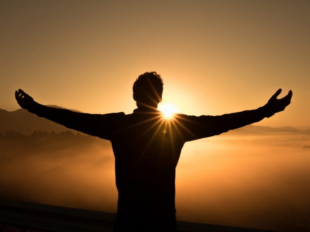 Silhouette of man with arms stretched out to the sides in front of a sunrise over misty mountains