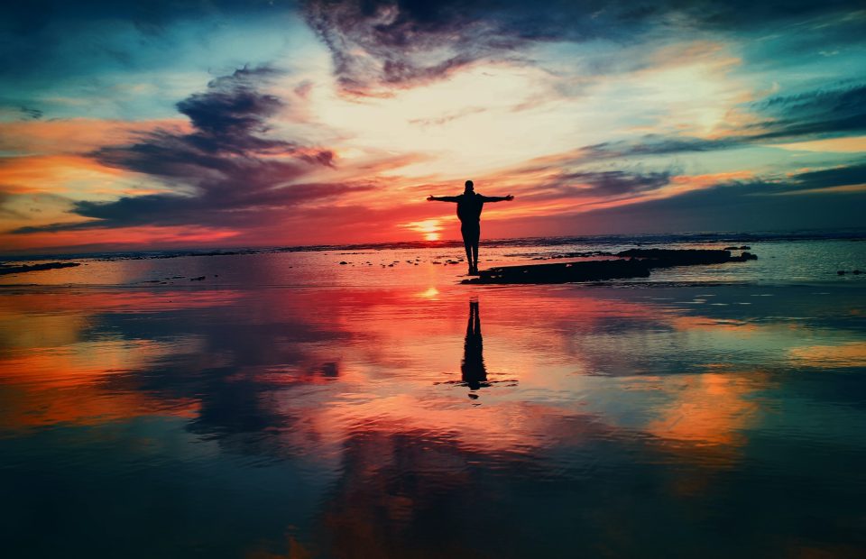 Person standing with arms wide open on a small reef with water all around and staring into a beautiful sunset