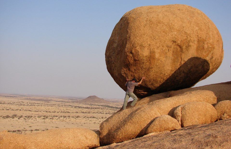 Desert scene with what appears to be a man holding back a gigantic boulder from falling down a rock pile