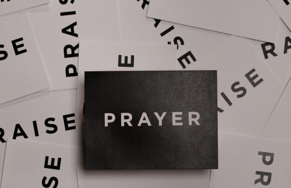 Black post card with the word Prayer in white centered on top of white post cards with the word praise scattered underneath